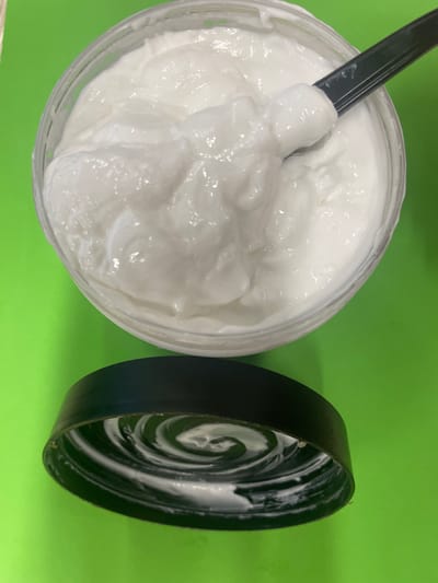 A close-up, top-down shot captures a clear plastic jar filled with a thick, white, creamy substance, possibly a cosmetic or food product. A dark green spatula is partially submerged in the cream, scooping a dollop of it. To the left of the jar, a black lid lies upside down, revealing a spiral pattern of the white cream adhering to its inner surface. The entire scene is set against a vibrant, uniform green background, suggesting an indoor setting rather than a specific outdoor location in the city of Терволово, Russia.
