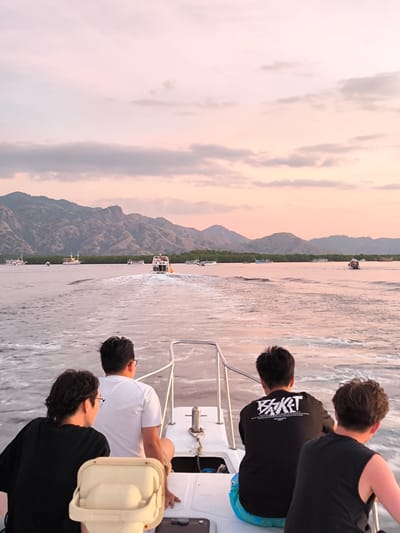 The image depicts a group of four young Asian men on a boat, traveling on the water. They are facing away from the camera, looking out at a scenic landscape. In the foreground, the boat's white railing and some equipment are visible. The men are casually dressed. The man on the far left wears a black t-shirt, the second man from the left wears a white t-shirt, the third man wears a black t-shirt with white text, and the man on the far right wears a black sleeveless t-shirt.
The background features a body of water with several other boats, some of which are carrying passengers. The water is slightly choppy, creating a wake behind their boat. Beyond the water, a line of green trees borders a range of imposing, rugged mountains. The sky above is a soft blend of pink, orange, and pale blue, indicating either sunrise or sunset. The overall atmosphere suggests a peaceful and scenic voyage. The location is identified as Rinca Village, Indonesia.