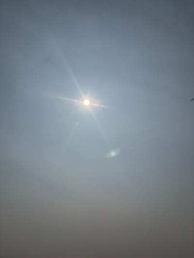 A bright sun is visible in the upper center of the frame, casting lens flares and visible light rays across a hazy, light gray sky. A smaller, green lens flare is located to the lower right of the sun. The bottom portion of the image is obscured by a dense, gray haze, making it impossible to discern any details of the environment or ground. The location is identified as Myandigi, Russia. There are no people or specific objects clearly visible.