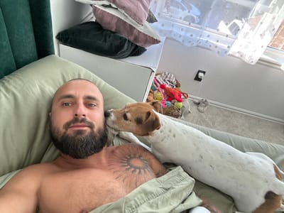 An overhead, eye-level shot shows a man lying in bed with his dog. The man has a shaved head, a beard, and is shirtless. He has a large sun tattoo on his chest. The dog, a Jack Russell Terrier, is white with brown markings and is licking the man's face. The man is lying on a light green pillow and blanket. Behind the man and to the left, there is a dark green headboard. To the right of the man, the dog's body extends out of the frame. In the background, there are stacked pillows of pink and dark green colors, a white box, a woven basket filled with dog toys, a window with sheer white curtains, and a wall with a white electrical outlet. The scene appears to be an indoor setting, likely a bedroom. The location is not specified within the city of Aurora, United States of America.
