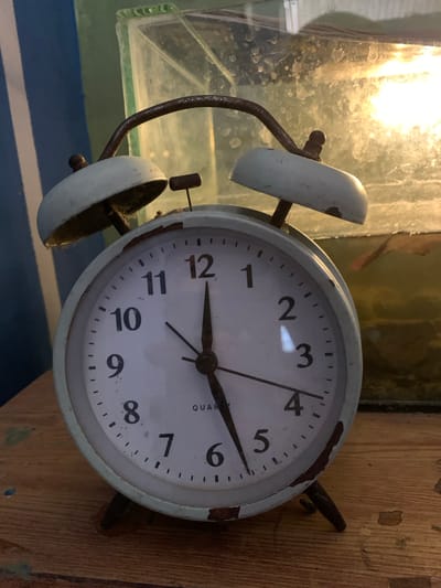 An aged, light blue alarm clock with a distressed finish rests on a wooden surface. The clock face displays Arabic numerals from 1 to 12, with a minute track. The hour hand points to the 12, and the minute hand points slightly past the 5. The alarm clock has two bells on top, connected by a curved metal handle. The background is out of focus, showing a glass container with what appears to be a light source behind it, possibly an aquarium or terrarium. The overall impression is one of an old, functional object in a domestic setting, with the specific location being the city of Karangkemiri, Indonesia.