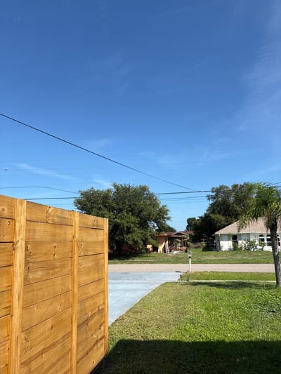 This image depicts a residential street scene on a clear, sunny day in North Port, USA. The perspective is from behind a newly constructed wooden fence, looking upwards and to the side. The bright blue sky dominates the upper left portion of the frame, with a few wispy clouds scattered across it. Several parallel power lines stretch across the sky, casting faint shadows.
To the right, the scene transitions to a well-maintained residential area. A paved driveway runs alongside a lush green lawn. In the background, houses with typical suburban architecture are visible, some with palm trees and other landscaping. One house in the mid-ground features a prominent A-frame roof and white siding, with arched windows. Another house, further back and to the left, has a more muted color scheme with visible foliage around it.
The most prominent object in the foreground is the wooden fence, constructed of vertical planks with horizontal support beams. Its warm, natural wood tone suggests it is recently built or stained. The overall atmosphere is peaceful and idyllic, characteristic of a suburban environment on a pleasant day. The time of day appears to be midday, given the bright sunlight and the quality of the shadows cast. No people or animals are visible, and there is no visible text in the image.
