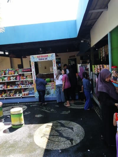 In the city of Tegalwangi, Indonesia, a street-side food stall named "ES TELLER TEKO 3R" is depicted under a blue overhang. The stall displays a variety of packaged snacks and drinks on shelves. A woman in a dark blue jacket and a patterned headscarf stands at the stall, interacting with the vendor. To her right, a woman in a white t-shirt and grey pants is visible. Several other individuals, including a child in a blue outfit, are gathered near the stall, some of whom are wearing hijabs. The ground in front of the stall is dark, with a decorative mosaic pattern of circles and fish-like shapes, and is partially illuminated by sunlight casting shadows. Two cylindrical containers, one green and yellow, and another light-colored one, are positioned on the left side of the foreground. The overall scene suggests a busy, open-air marketplace or recreational area.