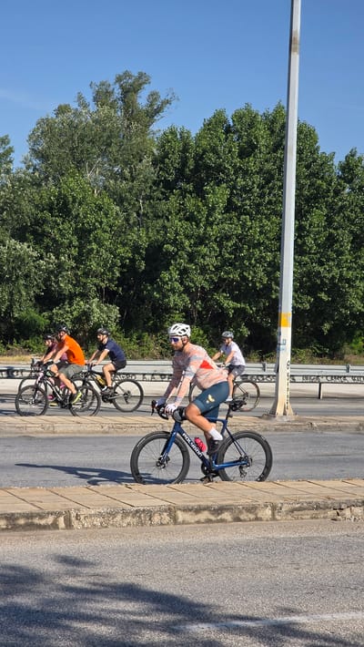 In Δημοτική Ενότητα Προμαχώνος, Greece, five individuals are seen cycling in a group on an asphalt road under a clear blue sky. The most prominent cyclist in the foreground is a male wearing a white helmet, sunglasses, a patterned long-sleeve cycling jersey, dark teal cycling shorts, white gloves, and white socks. He is actively pedaling a blue Polygon road bike with disc brakes and deep-rimmed black wheels.
Behind him, to the left, is another male cyclist in an orange t-shirt, dark shorts, and a helmet, riding a dark mountain bike or hybrid. Further back, a third male cyclist wears a dark blue t-shirt, dark shorts, and a helmet, also on a dark mountain bike or hybrid. Partially obscured to the left of the group, a fourth cyclist wears a purple top on a pink and black bicycle. The fifth cyclist, mostly obscured by a utility pole on the right, wears a white t-shirt, dark shorts, and a helmet.
The cyclists are positioned on an asphalt road with a concrete paver sidewalk to their immediate left, separated by a raised curb. Both the road and sidewalk exhibit signs of wear and cracking. In the background, a dense treeline of green foliage extends across the frame. A white metal guardrail runs horizontally between the road and the trees. On the right side of the road, a tall, grey, octagonal utility pole with yellow markings stands erect. The scene is brightly lit by natural sunlight, casting distinct, long shadows of the cyclists and the pole onto the road and sidewalk, indicating a low sun angle.