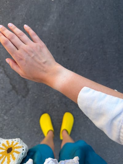 The image shows a first-person perspective of a person's arm and hand extended downwards towards the ground. The arm exhibits a distinct tan line on the forearm, suggesting sun exposure from a sleeve or bracelet. The person is wearing bright yellow slip-on shoes and turquoise shorts. A portion of a white knit garment with a yellow flower pattern is visible in the lower left corner. The background is a dark, textured asphalt surface, typical of a street or sidewalk. The scene is set outdoors. No specific city or location is identifiable from the image itself, and therefore, the mention of Xagħra, Malta is not supported by visual evidence.