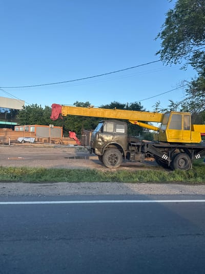 An older model, olive-drab truck with a yellow crane attached is parked on a gravel surface next to a paved road. The truck is facing right and the crane boom is extended over the cabin. The crane has a yellow cabin and a red hook at its end. In the background, there are stacks of lumber, an orange container, a building with reflective windows, and trees. The sky is clear and blue. The scene appears to be in an industrial or construction area in Vatra, Moldova.