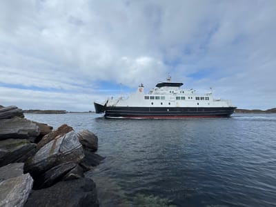 A ferry, identified by the name "LUROEY" and the number "8" on its funnel, sails on choppy blue water towards the right of the frame. The ferry is white with a black hull and a red waterline. The sky above is overcast with textured white clouds. In the foreground, a pile of large, grey, and brown rocks juts into the water from the left side of the image. In the background, a small, treeless island with a few scattered red buildings is visible. The scene appears to be a coastal area in Lovund, Norway. There are no people visible in the frame.