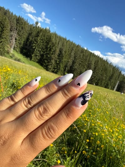 A close-up shot captures a person's hand with intricately designed fingernails in the foreground, set against a blurred backdrop of a mountain meadow in Yundola, Bulgaria. The nails are long and pointed, painted in a matte white finish. Several nails feature small, black heart decals. One nail, at the forefront, is adorned with a detailed black and white image of Mickey Mouse's face, complete with his signature ears and eyes. The hand is illuminated by bright sunlight, casting subtle shadows on the skin.
The background depicts a vibrant green meadow dotted with yellow wildflowers, indicative of a summer bloom. Beyond the meadow, a dense forest of evergreen trees covers the slope of a mountain. The sky above is a clear, bright blue with scattered white, fluffy clouds. The overall scene conveys a sense of natural beauty and a touch of playful artistry.