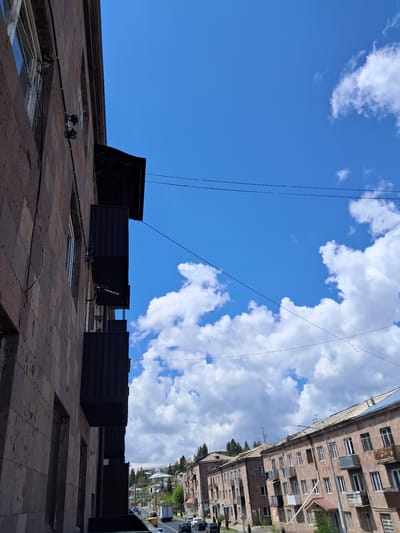 The image, captured in Gavar, Armenia, presents an upward-looking view of an urban landscape under a bright blue sky adorned with scattered white, fluffy clouds. The scene is bisected by the horizon line of rooftops.
In the foreground, a large, multi-story building constructed of reddish-brown stone or brick rises prominently. Its upper facade and rooftop are visible, featuring several windows or skylights, and a series of dark, rectangular structures that appear to be utility boxes or ventilation units. Two small, dark objects resembling spotlights or security cameras are mounted on a ledge on this building. Multiple thin, dark utility lines stretch across the sky, originating from or passing over these buildings.
Beyond the immediate foreground building, a street or open area is visible, separating it from a row of distant residential buildings. These background structures are multi-storied and appear to be made of a similar brown or beige-toned material, characterized by numerous windows and some enclosed balconies, typical of apartment blocks. Some green trees are interspersed among these buildings, adding a touch of greenery to the urban setting.
The time of day is clearly daytime, with bright sunlight illuminating the scene and casting clear, soft shadows. The weather is fair, with a predominantly blue sky and well-defined clouds. There are no visible people or specific activities taking place, and no discernible text is present in the image. The architectural style of the buildings is consistent with urban environments in the region, reflecting the location in Gavar, Armenia.
