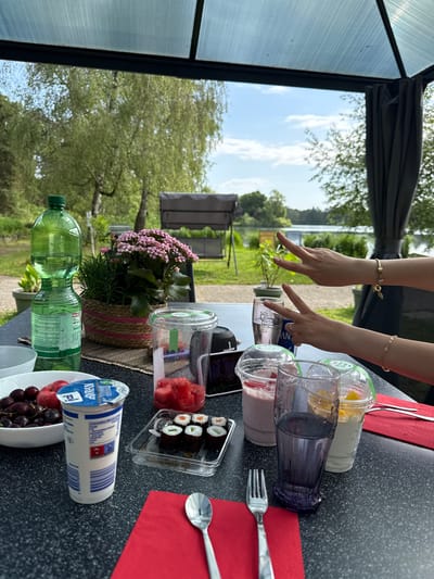 The image shows a table set with various food and beverages, seemingly outdoors with a view of a lake and greenery, possibly in Hanau, Germany. On the table are smoothies, a cup of yogurt, a container of sushi, a bowl of cherries, and bottles of water. There are also red napkins, a spoon, and a fork. A person's hands are visible, positioned over the items. The scene appears to be a casual outdoor meal or gathering. The presence of trees, grass, and a glimpse of water indicates a lakeside or park setting. The structures in the background suggest a park or recreational area.