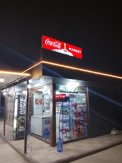 The image depicts a small market at night in Sabir Rakhimov, Uzbekistan. The building has a Coca-Cola Market sign on top. The market sells Gorilla energy drinks and hot dogs. A Pepsi-branded freezer is inside, and several plastic water jugs are nearby. The market is constructed with glass walls on three sides, allowing a view of the interior merchandise. The area around the market is paved. String lights are hung along the roofline.