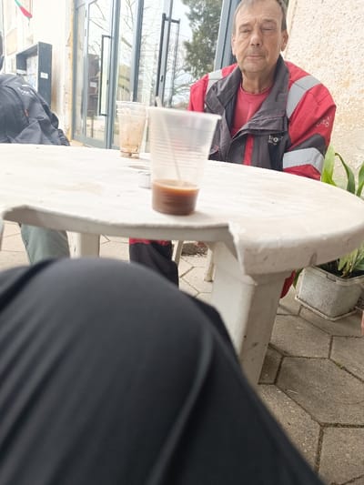 An outdoor scene in Vinitsa, Bulgaria, depicts a low-angle perspective of an informal gathering. A middle-aged male individual with short brown hair and a serious expression is seated at a white, worn plastic outdoor table, looking directly at the camera. He wears a red t-shirt under a black and red jacket with grey reflective accents and "BOSC" branding on the left chest. On the table in front of him, two clear plastic cups are present; one contains a small amount of dark brown liquid and a stir stick, while the other is mostly empty. A small, silver object is next to the front cup. The legs of two other individuals are partially visible at the table: the photographer's dark-clad legs in the foreground, and another person's leg in green pants on the far left. The ground is paved with hexagonal or octagonal stones. In the background, a light-colored building facade with large glass double doors reflects trees and an overcast sky. A dark wall-mounted shelf is visible on the far left. A potted plant in a white rectangular planter sits on the ground to the right of the table.