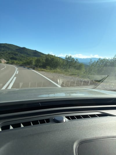 The image depicts a view from inside a vehicle, looking out through the windshield onto a winding road that travels through a mountainous, forested landscape under a clear blue sky. The road has white lane markings. The dashboard of the vehicle is visible in the foreground, with air vents and a speaker grill. In the distance, mountains and trees are visible. The scene is likely located in or near Ponikvica, Montenegro, given the geographical context suggested by the terrain and the request. There are no people or animals visible in the image, nor any discernible action or interaction. The environment is natural and rural.
