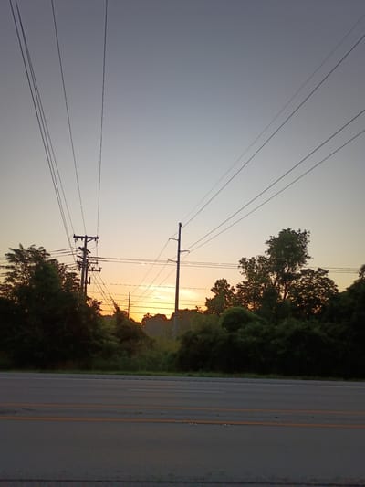 The image depicts an outdoor scene at dawn or dusk, with a clear sky transitioning from a pale blue at the top to a soft orange hue near the horizon. Several power lines stretch across the frame, converging towards telephone poles that stand amongst dense green foliage. In the foreground, a paved road with double yellow lines is visible, suggesting a highway or a major street. The overall impression is one of a rural or suburban setting, possibly in Frankfort, USA, where the natural beauty of the sky meets the infrastructure of civilization.