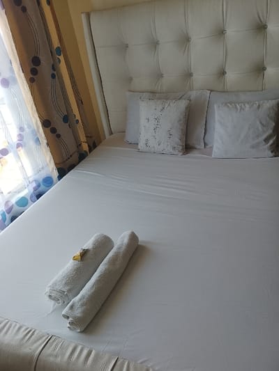 This image depicts a bedroom interior located in Gatongora ward, Kenya. The central subject is a large bed with a prominent white, tufted headboard featuring multiple button accents. The bed is neatly made with white sheets. Four pillows are arranged against the headboard: two standard white pillows, one white throw pillow with a subtle textured or patterned design, and another plain white throw pillow. At the foot of the bed, two white towels are rolled and placed side-by-side. A small, gold-colored, wrapped item rests on the leftmost rolled towel. To the left of the bed, a window is partially obscured by a sheer curtain adorned with blue, purple, and gray circular and wavy patterns. A darker, opaque curtain in brown/gold tones is visible behind the sheer curtain. The walls are a light, neutral color. No individuals or active interactions are present within the scene.