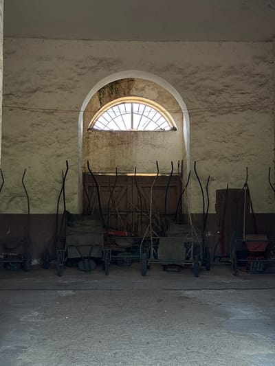 The photograph shows an interior scene in Слобода, Russia. A line of wheelbarrows is parked in front of a weathered wall with an arched window. The window, featuring a grid pattern, admits light into the otherwise dimly lit space. The wall texture is rough and uneven. The wheelbarrows are of varying designs, some with visible framework and others with covered compartments. The floor appears to be concrete.