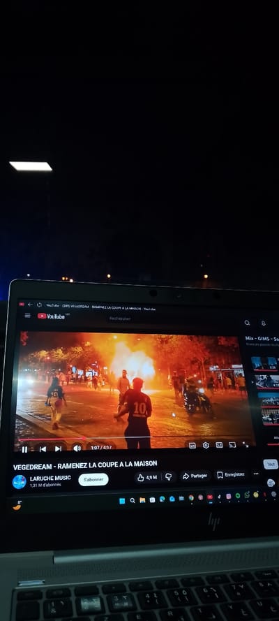 A laptop displays a YouTube video titled "VEGEDREAM - RAMENEZ LA COUPE A LA MAISON." The video shows a street scene with a burning object, possibly a vehicle, with people nearby. Two figures wearing football jerseys are prominent in the foreground. The YouTube interface is visible, including playback controls, channel information ("LARUCHE MUSIC"), and various icons for liking, sharing, and saving the video. The laptop's keyboard is also in view.