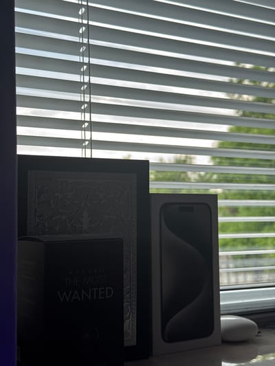 The image presents an indoor still life scene, likely on a shelf or windowsill, in Kardam, Bulgaria. A window on the left side is covered by vertical white or light grey blinds, through which bright daylight enters, creating distinct shadows and highlights. Beyond the blinds, a blurred view of green trees or bushes is visible, indicating an outdoor natural setting.
To the right, several objects are arranged. A light-colored box, strongly resembling smartphone packaging, specifically an iPhone box, is partially visible at the top, displaying the silhouette of a device's camera module. Below this, a dark, rectangular decorative item, possibly a frame or a box, features an intricate, ornate pattern. Resting against or in front of this patterned object is a black fragrance box. The visible text on this box clearly reads "AZZARO THE MOST WANTED" in a light-colored font. Below it, another section of a box also shows "AZZARO". On the far right, a small, smooth, white decorative object, possibly made of ceramic, is partly visible against the wall or window frame. The strong contrast between the bright light from the window and the darker interior suggests it is daytime.