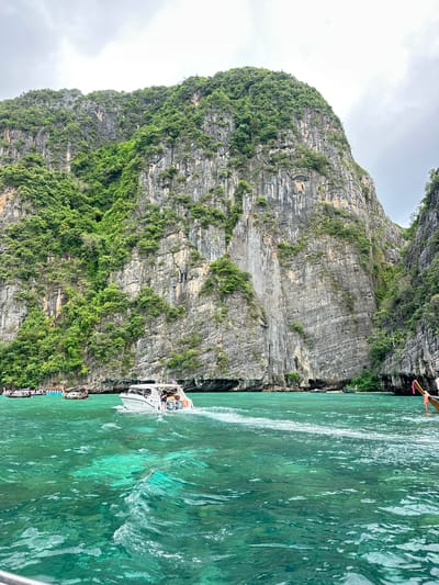 The image depicts a view of the ocean and a towering, vegetated limestone cliff face. The water is a clear, vibrant turquoise green, with visible ripples and a white wake trailing from a white motorboat in the foreground. The motorboat, carrying several passengers, is heading away from the viewer and towards the left of the frame. In the distance, other boats, including what appear to be traditional longtail boats, are moored near the shore. The cliff face, a dominant feature of the image, is grey and textured, with lush green vegetation clinging to its slopes and crevices. The sky above is overcast with grey clouds. The scene is situated in Haad Yao, Thailand, a location known for its stunning natural beauty, particularly its dramatic karst topography and clear waters.