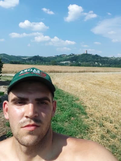 This is a selfie of a young man wearing a green baseball cap with red text. The man is shirtless, and the sun is reflecting off his face, suggesting it is a warm day. He has short brown hair and a light beard.
Behind the man, the scene is a rural landscape under a bright blue sky with scattered white clouds. The immediate foreground features a field of dry, golden stubble, indicative of a harvested crop, possibly wheat or barley. A strip of green grass runs along the left edge of this field.
In the middle ground, rolling hills covered in lush green trees rise up. Perched atop one of these hills is a cluster of buildings and a tall, reddish tower, which is a prominent landmark. The overall setting appears to be in a hilly, agricultural region.
The text on the cap is partially visible and appears to read "SAGRA" in large letters, with smaller text below. The location context identifies this as Guarene, Italy. The bright sunlight and clear sky suggest it is daytime, likely late morning or afternoon, during a warm season. The man's expression is neutral, and the overall mood is relaxed and casual, typical of an outdoor selfie.
