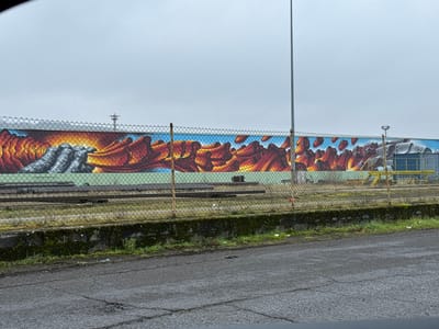 This image depicts an outdoor industrial or former industrial scene in Aubange, Belgium, under an overcast, light grey sky. In the foreground, a dark grey asphalt surface, showing cracks and minor wear, occupies the bottom portion. Above the asphalt, a chain-link fence, supported by metal posts with visible rust, extends horizontally across the frame. The base of the fence rests on a low concrete retaining wall covered with moss and sparse dry grass. Behind the fence, several parallel disused railroad tracks are visible, spanning the width of the image over a ground of dirt and dry vegetation.
The main subject is a large, colorful mural covering a long, solid wall in the background. The mural features stylized, abstract shapes. On the left side, grey, rock-like or solidified forms transition into vibrant orange, red, and yellow masses, resembling molten material or fire against a light green base. Towards the right, similar fiery-hued shapes appear suspended or floating against a blue background, suggesting a sky. A tall, slender metal light pole stands in the mid-right section of the scene, partially obscuring the mural. Further to the right, behind the fence, a blue industrial structure or building is visible, with yellow support beams or equipment in front of it. In the far distance, above the mural wall, a metal tower or antenna structure is faintly discernible against the sky. No individuals are present in the scene.