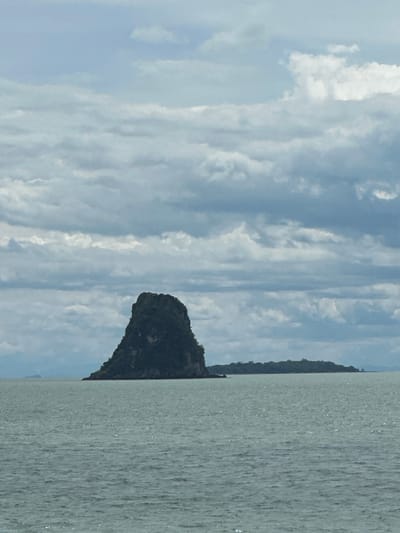 The image shows a body of water with several islands in the distance. The sky is cloudy and overcast. The water appears to be choppy, with small waves. The islands are covered in lush green vegetation. In the foreground, the water is a muted green color. In the mid-ground, a larger island with a prominent rock formation rises out of the water. To the right of this island, a smaller, flatter island is visible. The sky is filled with a layer of gray and white clouds. The overall impression is one of a natural, serene landscape.