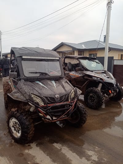 The scene depicts two muddy CFMOTO UTVs parked on a wet, paved road in Stary Chultem, Russia. The UTV on the left is predominantly black with red and orange accents, while the one on the right has orange stripes. Both vehicles are covered in mud splatters. In the background, a brick house with a gray tiled roof is visible behind a metal fence. A utility pole with power lines extends overhead. People are standing near the left UTV. The sky is overcast.