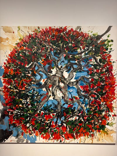 This is an abstract painting of a tree with vibrant red and green leaves, set against a blue sky. The tree's trunk and branches are depicted with thick, dark brushstrokes, giving them a sense of texture and movement. The leaves are rendered in a more impressionistic style, with dabs of color that suggest their fullness and vibrancy. Splashes of paint in various colors, including white, yellow, and brown, are scattered across the canvas, adding to the abstract and energetic feel of the artwork.
The painting evokes a sense of nature's beauty and the raw power of the elements. The bright colors and dynamic brushwork suggest a lively and perhaps windy atmosphere. The composition, with the tree filling most of the frame and branches reaching towards the viewer, creates an immersive experience. The interplay of light and shadow, though abstractly represented, adds depth and dimension to the scene.
The setting appears to be outdoors, given the depiction of the tree and sky. The context of La Cuesta, Spain, might suggest a Mediterranean landscape, known for its sunny weather and lush vegetation. However, the abstract nature of the painting allows for a more universal interpretation, focusing on the essence of the tree and its natural surroundings.
There is no visible text in the image. The overall emotion conveyed is one of vitality, dynamism, and the untamed beauty of nature.