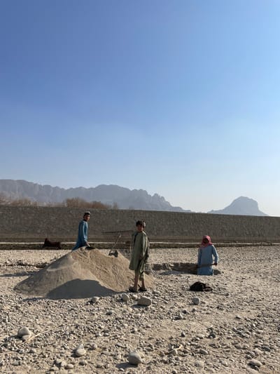 The image depicts a dry, rocky terrain in Shuyene Vosta, Afghanistan, under a clear blue sky, with distant mountains forming the horizon. Three individuals, appearing to be young males, are present. On the left, one individual in a light blue tunic stands atop a large pile of light-colored sand. Another individual, in an olive-tan tunic, stands to the right of the sand pile, holding a shovel with its blade resting on the sand. A third individual, wearing a light blue tunic and a reddish head covering, is partially submerged in a rectangular pit in the ground on the right, holding a shovel. A brown saddle rests against a low stone wall in the mid-ground, behind the left-most individual. Another dark fabric object lies on the ground near the pit on the right. The ground is covered with gravel and scattered larger stones.