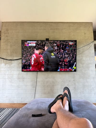 An interior scene captured from a low perspective shows a person's lower legs and feet, clad in black flip-flops, resting on a grey upholstered surface in the foreground. A black remote control lies on the upholstery to the left of the feet. In the midground, a large, flat-screen television is mounted on a textured concrete wall featuring horizontal grooves. The television displays a soccer match in progress. The on-screen graphic in the upper left corner indicates "MUN 0 - LIV 7" at 90:57, with a "Substitution" notification showing "8 Fernandes" out and "23 Shaw" in. The upper right corner of the screen displays "ESPN VIVO". The television image features a blurred player in a red jersey with the number "8" on the back, facing a man in a black puffer jacket with a yellow logo. A stadium crowd is visible in the background of the television screen. Black electrical cables are visibly run along and mounted on the concrete wall around the television. A small black device, possibly a camera or sensor, is positioned on top of the television. The wall above the television transitions to a smooth, light-colored surface. A striped rug and a light-colored wooden floor element are partially visible on the floor in the bottom left of the image. The specific city of Santo Domingo, Chile, is not verifiable from the visual information provided.