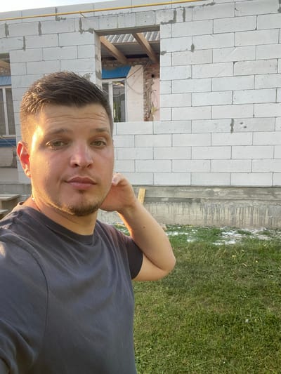The image shows a man posing for a selfie outdoors. He is wearing a dark grey t-shirt and has short, dark hair and stubble. The man has green eyes and a neutral expression. He is resting his left hand on the back of his neck.

The setting appears to be a construction site. To the left of the man, there is a wall made of white cinder blocks, with an unfinished opening revealing wooden beams and some exposed brickwork inside. To the right, there is a patch of green grass and what looks like a concrete foundation with vertical grooves. The lighting suggests it is late afternoon, with the sun casting a warm glow on the man's face and hair.

There is no visible text in the image. The overall mood is relaxed and casual, with the man appearing comfortable in his surroundings. The location context is Yablunivka, Ukraine.