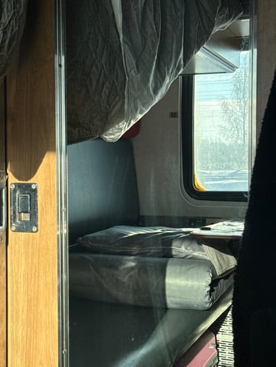 The image displays the interior of a train compartment, situated in Volkhov, Russia, viewed towards a window during a bright, sunny winter day. On the left, a light brown wooden panel, possibly part of a door frame or partition, features a dark, weathered metallic latch mechanism and is bordered by a chrome trim. Strong sunlight casts distinct shadows on this wooden surface.

In the central part of the frame, a grey quilted blanket or bedspread hangs from an unseen upper bunk. Below it, a grey upholstered seat or lower bunk is made up with a folded grey sleeping pad and a grey pillow, both partially illuminated by sunlight. Behind this sleeping area is a darker grey upholstered backrest. A small, light-colored wooden table is positioned under the window, holding what appears to be folded fabric or paper. Below the table, a power outlet or control panel is visible. A white rectangular panel marked "32" is affixed to the wall above the seat, near the window. A red suitcase is partially visible beneath the lower bunk. The far right foreground is partially obscured by a dark, vertical fabric element.

Through the train window, a clear winter landscape is visible, featuring bare trees, possibly birches, against a bright, clear blue sky. The ground outside is covered in snow, and a distant treeline is discernible. No individuals are present, and no actions are depicted within the scene.
