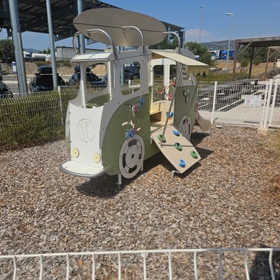 This is a daytime, eye-level, outdoor, full-shot photograph of a children's playground structure designed to resemble a vintage camper van. The structure is primarily white and green, with a light brown awning extending from its side. A small, angled climbing board with blue and green handholds is attached to the side of the van. The playground is situated on a bed of wood chips, and a white metal fence runs across the foreground. In the background, several cars, including two black SUVs with roof racks, are parked under a solar panel-covered structure. Beyond the cars, there are trees, hills, and a clear blue sky. A sign with international symbols is visible on the fence. This playground is located at a rest stop on the A9 motorway in La Palme, France.