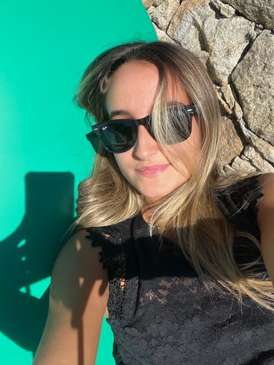 A female subject is depicted in a close-up, head-and-shoulders portrait, consistent with a selfie, situated in the city of Oliveira de Frades, Portugal. She has long, wavy, blonde hair and wears dark Ray-Ban Wayfarer-style sunglasses and a black lace top with cap sleeves. Her lips are slightly parted, indicating a faint smile, and she looks directly at the camera. Strong, direct sunlight illuminates her from the upper left, casting a distinct shadow of an unidentified object, consistent with a smartphone, on the vibrant green surface to her lower left. The background is divided, with a solid green surface occupying the left portion and a rough-hewn stone wall, composed of irregular grey and beige stones, forming the right portion.