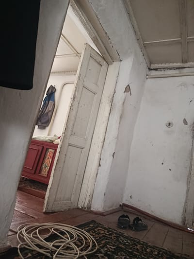 This image depicts an interior scene, likely in Yusupov, Uzbekistan, characterized by aged architecture and signs of wear. The primary subjects are the interior of a room with a doorway, revealing another space beyond. Visible objects include a red cabinet with a decorative panel, a hanging jacket, a coil of white rope on a patterned rug, and a pair of dark sandals on the floor.

The scene appears to be set indoors, in what looks like a room with wooden floors and white, textured walls that show some damage and peeling paint. The doorway is partially open, revealing an interior with a white ceiling that has exposed wooden beams. The lighting suggests it could be daytime, but there are no strong indicators of specific time or weather conditions.

Notable details include the rustic nature of the building, with its aged wooden door and textured walls. The arrangement of the objects, such as the coiled rope and the sandals, suggests a lived-in space. The presence of traditional-looking decor on the red cabinet might hint at a cultural context. There is no visible text in the image. The overall impression is one of a simple, lived-in dwelling that has seen better days.