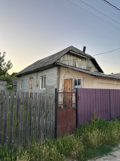 This image depicts an exterior view of a rustic, weathered house with a pitched roof covered in gray shingles. The house is constructed with a mix of materials, including what appears to be rough concrete on one section and aged, painted wooden planks on another. A dark purple corrugated metal sheet forms part of the upper wall. There's a wooden fence in the foreground, with some of the planks showing signs of wear and graying. Overgrown green grass with feathery seed heads grows alongside the fence and up against the metal sheeting.
Visible objects include a metal gate with decorative scrollwork and a simple metal door. Several windows are present; one has what looks like a boarded-up or covered opening. Electrical wires are strung across the sky, and a small chimney protrudes from the roof. The lighting suggests it is either early morning or late afternoon, with the sky displaying a soft gradient of blue and yellow. The overall impression is one of a rural, possibly aged property, with elements of disrepair and a natural, overgrown surrounding. No people or animals are visible. The location is identified as Pîrlița, Moldova.