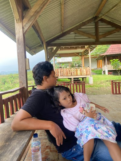 A man and a child are resting in a structure in Penanggungan, Indonesia. The man is wearing a black t-shirt and blue jeans. The child, who appears to be a young girl, is wearing a light pink shirt with a Minnie Mouse graphic and a floral patterned skirt. The child is lying on the man, holding a cellphone with a decorated case. They are in a wooden structure with an exposed roof frame. A plastic bottle of water is on the table beside them. Outside the structure, there is a view of rice terraces and vegetation. A building is visible in the background, and there is a grassy area.