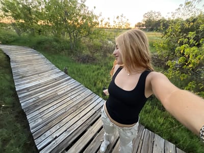 A woman in a black tank top and light-colored pants stands on a wooden boardwalk, taking a selfie. The boardwalk is surrounded by green grass, shrubs, and trees, characteristic of the Khwai region in Botswana. The lighting suggests either sunrise or sunset. The woman is wearing a necklace and bracelet.