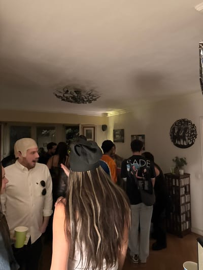 A gathering of people indoors, likely a party. The room is lit dimly. Several individuals are visible, including one wearing a white wig and a white shirt with sunglasses pinned to it, holding a green cup. Another person wears a backward baseball cap. Others are standing in groups further down the room. Wall decorations include metal sculptures and framed pictures. A dark cabinet sits against the far wall.