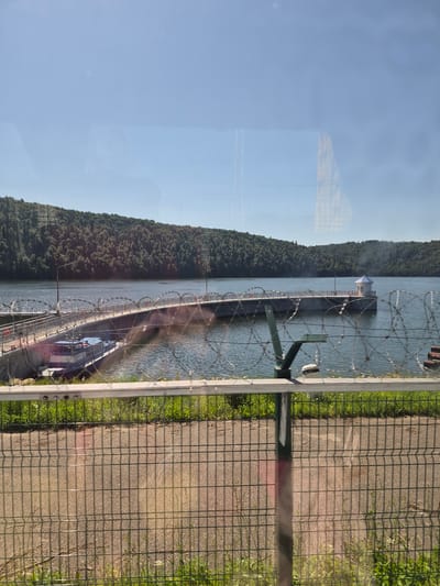 The image shows a view of a reservoir with a pier extending into the water. The pier has a structure at its end and is equipped with railings and barbed wire. A small boat is docked at the beginning of the pier. The reservoir is surrounded by a forested hillside. In the foreground, there is a wire fence and some grass. The sky is clear and blue. The location is identified as Pavlovka, Russia.