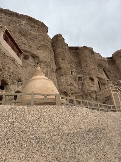 The image depicts the Yulin Caves, located in Guazhou County, Gansu Province, China. The caves are carved into the sandstone cliffs. Visible are cave entrances with ventilation grates. A stupa-like structure is situated in front of one cave. A modern metal and concrete staircase or walkway runs alongside the cliff face, providing access to the caves. The sky is overcast.