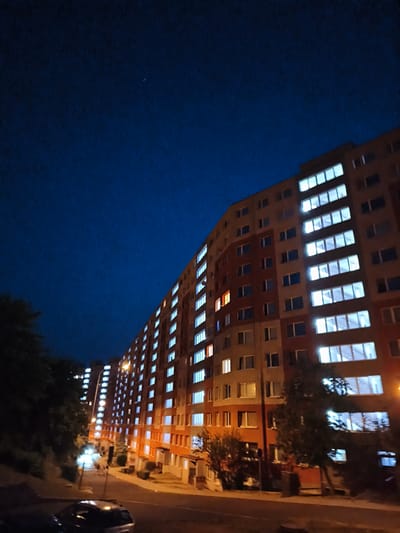 The image shows a nighttime view of a large apartment building, likely in the city of Most, Czechia. The building is illuminated by the lights from its many windows, with a few windows showing a warm, orange glow, possibly from interior lights. The sky above is a deep, dark blue. In the foreground, there is a street with at least two cars parked. Trees and bushes are visible on the left side of the image, and a street lamp illuminates the area. The perspective is from a slightly elevated viewpoint, looking up at the building and across the street.