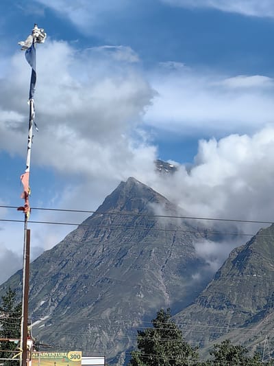 In Yamling, India, a flagpole stands tall against a backdrop of a majestic mountain range, partially obscured by billowing clouds. The flagpole, adorned with torn prayer flags, juts into the blue sky, a stark contrast to the rugged, green slopes of the mountains behind. Power lines crisscross the scene, hinting at civilization amidst the natural grandeur. The lower portion of the image reveals a sign, partially visible, that reads "AT ADVENTURE" and displays "Counter No 300," suggesting a tourist or recreational area. The overall scene is one of serene beauty, where human elements blend with the awe-inspiring power of nature.