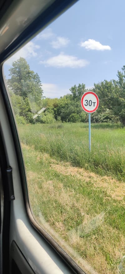 The image was taken from inside a vehicle, looking out the window. A traffic sign with "30т" in white text on a red circle is visible in the foreground, mounted on a blue pole. The sign indicates a weight limit of 30 tons. The background consists of a grassy field with scattered trees and a bright blue sky with some white clouds. The scene is set in Kolomak, Ukraine, as indicated by the location. The image does not contain any people or actions.