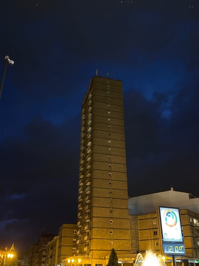 Here is a description of the image.
The image captures a tall, rectangular building under a dark blue evening sky, likely taken at night. The building is light brown or beige, with a repetitive pattern of small windows and horizontal lines suggesting multiple stories. To the right of the building, a large billboard displays what appears to be a colorful logo and some digital text, including the number "21:06". Streetlights and other buildings are visible in the background, indicating an urban setting. A security camera can also be seen in the bottom left corner.
Based on the presence of the San Lázaro building and the billboard, the location can be identified as Oviedo, Spain. Specifically, the tall building is the Residencial San Lázaro. The billboard is near the intersection of Avenida de Colón and Calle Samuel Sánchez, indicating the image was taken from that vicinity.