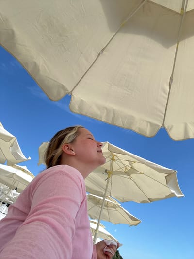 A young person with blonde hair, wearing a pink top, is seen from the side, looking up with their eyes closed, as if enjoying the sun. They are under several beige beach umbrellas, which are positioned against a clear blue sky. The umbrellas are arranged in rows, suggesting a beachside setting. The perspective is from below, looking up at the person and the umbrellas. The background is a bright blue sky with a few subtle cloud formations. The scene appears to be a warm, sunny day, likely at a beach resort in Lermontovo, Russia, given the presence of beach umbrellas and the clear sky.