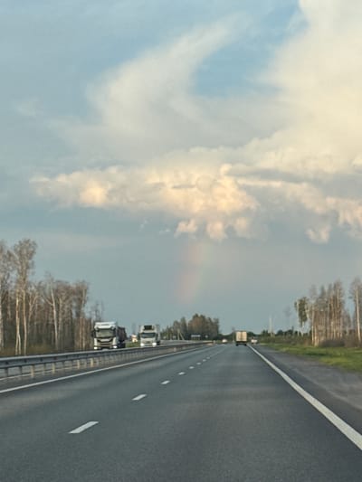 A view from the perspective of a vehicle traveling on a highway in or near Bogdanovich, Russia. The road has multiple lanes marked by white lines, and a metal guardrail runs along the left side. Trees, mostly bare of leaves, line the side of the road. In the distance, several trucks are visible on the highway. The sky above is partly cloudy with a faint rainbow appearing in the distance. The lighting suggests it is either early morning or late afternoon.