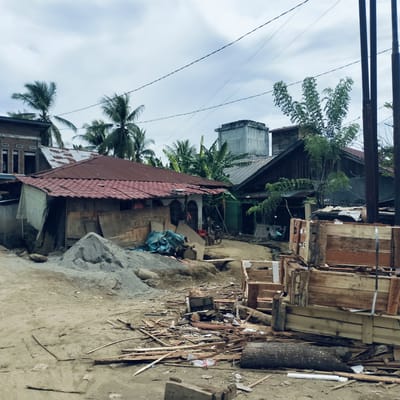 This outdoor scene, set in Beurawang, Indonesia, depicts a bustling residential area under an overcast sky. The foreground is dominated by unpaved, dusty ground littered with various pieces of wood, construction debris, and a large fallen log. On the left, a substantial pile of sand or gravel suggests ongoing construction or renovation, with blue tarps or bags nearby.

Several houses and structures are visible throughout the image. To the left, a single-story dwelling with a prominent red tiled roof appears somewhat makeshift, with plywood and other materials leaning against its white-washed walls. Behind it, a brick building with visible window frames is partially obscured. Further into the mid-ground, a dark, wooden-sided house with a dark roof stands, and further back and to the right, a tall, grey concrete structure, possibly a water tower or a swiftlet house, rises above the rooftops.

Lush tropical vegetation is abundant, with several tall palm trees (likely coconut palms) dotting the landscape, especially towards the left and center-background. Banana plants are also visible behind some of the houses. Electrical wires crisscross the sky, connecting to several tall, dark utility poles on the far right of the frame.

A bicycle is parked near the red-roofed house, and a small scooter or motorcycle is visible further back on a dirt path. Among the scattered debris in the foreground, wooden crates or pallets are stacked, adding to the impression of a working or developing area. A red Coca-Cola crate is visible near the bicycle. The scene captures a typical daytime view of a community in a tropical setting, characterized by mixed housing styles, natural elements, and everyday clutter. No specific human activity is clearly depicted, suggesting a quiet moment in a lived-in environment.