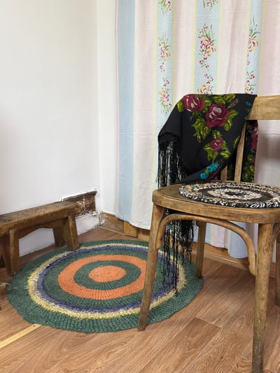 In a dimly lit room, likely in Ust'-Pyzas, Russia, a rustic wooden chair stands as the focal point. A black shawl with a vibrant floral pattern of red roses and green leaves is draped over its back. The shawl is edged with black fringe that hangs down. On the seat of the chair rests a small, circular braided rug, predominantly black and white with brown accents. Beside the chair, a simple wooden stool, worn and aged, is positioned near a wall. A circular rag rug with concentric rings of green, orange, purple, and yellow lies on the wooden floor. The wall behind the objects is painted white, and a set of pastel-colored curtains, featuring stripes and floral motifs, hangs to the right. The floor is made of wooden planks. The scene evokes a sense of traditional rural living.