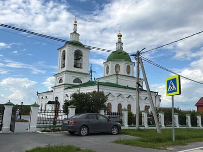 The image captures a white church with green domes and crosses, a bell tower, and a main dome, situated in the city of Kamenka, Russia. A dark gray sedan is parked on the side of a paved road in front of the church's white fence, which has ornate black metal railings and white pillars topped with green accents. A pedestrian crossing sign is visible to the right of the church. In the background, a person wearing a white shirt and pants is standing near the entrance of the church grounds, which are enclosed by a white gate. The sky is partly cloudy with a mix of blue sky and white clouds. Electrical wires are strung across the upper part of the image.